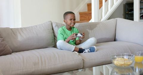 Young boy playing video game on living room couch with wireless controller and snacks