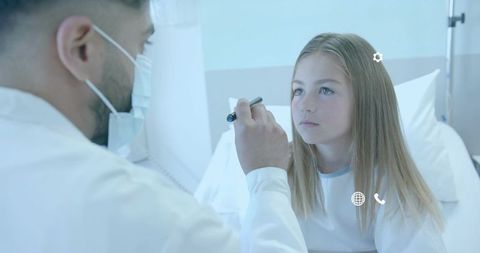 Pediatric eye exam using penlight on young girl in hospital bed by masked doctor