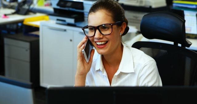 A young Caucasian woman is engaged in a conversation over the phone, smiling as she works at her office desk, with copy space. Her professional demeanor suggests she could be a customer service representative or an office administrator.