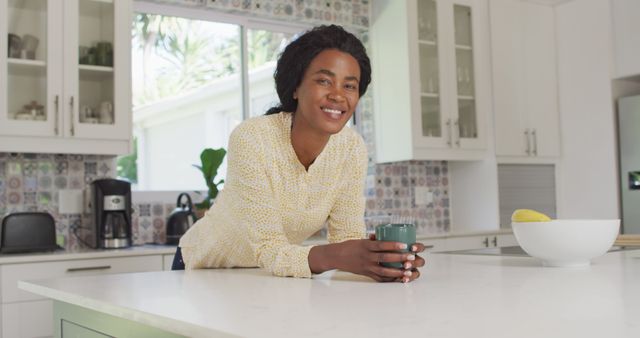 Woman leaning on kitchen island holding a coffee cup and smiling. Modern kitchen with white cabinets and patterned backsplash. Great for content on morning routines, home life, and lifestyle.