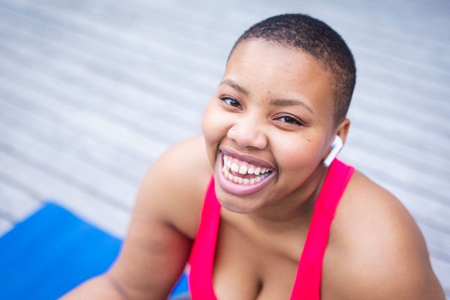 This image shows an African American woman with short hair smiling while sitting on a yoga mat outdoors. She is wearing casual fitness attire and earbuds, suggesting she might be listening to music or a podcast. This image can be used for promoting fitness, healthy lifestyle, wellness programs, outdoor activities, and mental well-being.