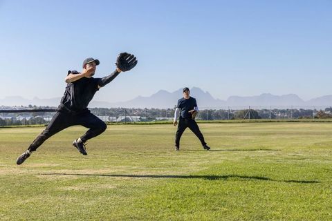 Mature players leaping to catch baseball in sunlit grassy outfield with mountain backdrop