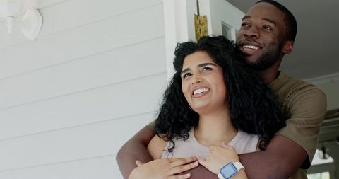 Sunlit diverse couple embracing on porch, man hugging woman from behind, smiling together