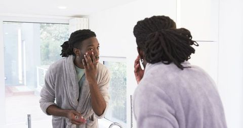African American man applying eye cream at vanity mirror wearing gray bathrobe