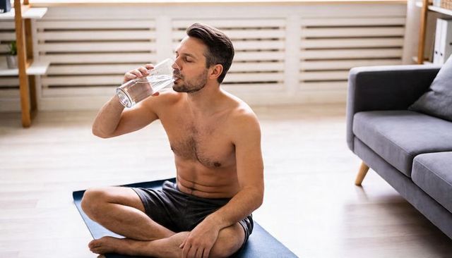 Shirtless man drinking water after home yoga session on blue mat