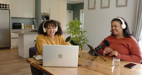 Diverse women recording cozy home podcast at kitchen table with laptops and microphones