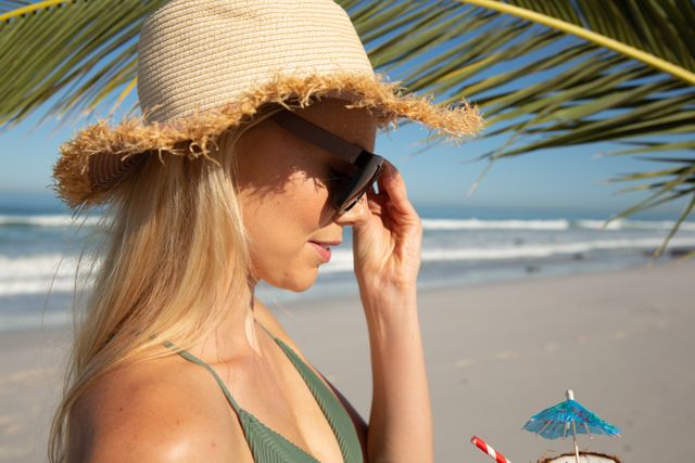 Caucasian woman enjoying time at the beach on a sunny day, wearing sun hat and sunglasses with palm tree sea in the background. Holiday relaxing summer wellbeing.
