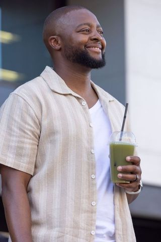 Smiling man holding green smoothie wearing casual beige shirt outdoors
