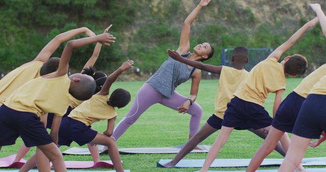 Fitness instructor guiding children in outdoor yoga poses on grass. Kids wearing athletic clothing following instructor's movements, emphasizing physical activity and teamwork. Ideal for use in fitness promotions, educational materials, and healthy lifestyle campaigns.