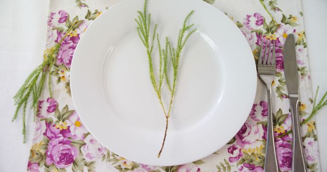A white plate is adorned with a simple plant stem, set on a floral tablecloth, with cutlery on the side, with copy space. This setting suggests a fresh, springtime meal or a special occasion like a garden party or Easter brunch.