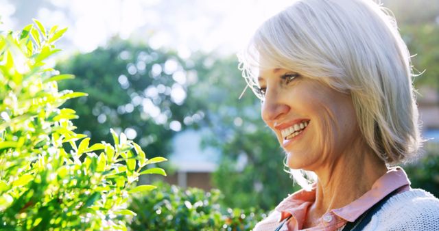 Senior woman smiling and enjoying nature in a sunny, lush garden. This image can be used for articles on healthy aging, outdoor activities for seniors, gardening, lifestyle, wellness, mental health, promoting happiness and relaxation for elderly individuals.