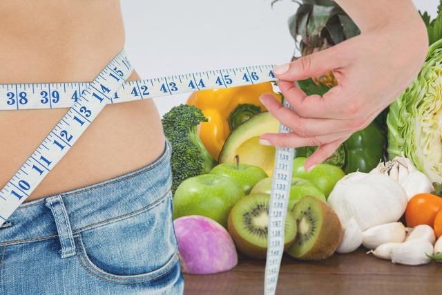 Midsection of a woman measuring her waist using a tape measure, surrounded by an assortment of fresh vegetables and fruits in the background. Emphasizes the importance of healthy eating and fitness in weight loss journeys. Ideal for use in health and wellness blogs, dietary websites, fitness programs, and social media promotions focused on nutrition and healthy lifestyle choices.