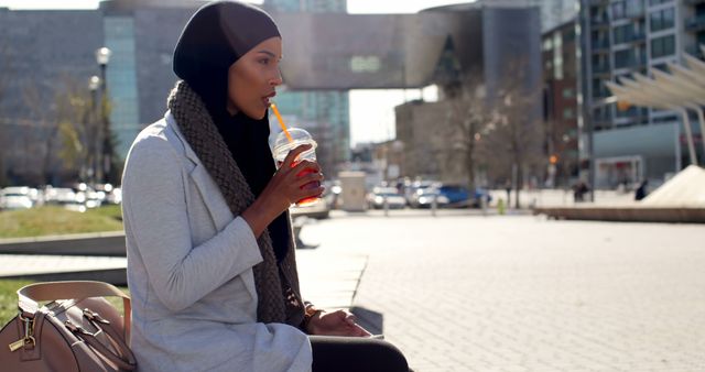 Woman wearing hijab drinks from a cup with a straw while sitting in a cityscape. Great for depicting urban lifestyle, cultural diversity, women's lifestyle, and outdoor activities. Use for articles on modern urban living, diversity and inclusion, or lifestyle blogs.