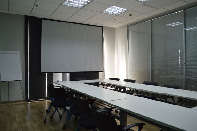 Bright modern meeting room shown with conference tables arranged around an empty projection screen. Flip chart placed at the corner. Benching style chairs with blue accents, emphasizing professionalism. Use for themes around business meetings, corporate settings, office environments, workspace design, and professional presentations.