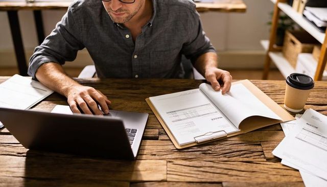 Freelancer reviewing invoices while typing on laptop at rustic wooden home office desk