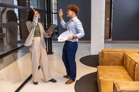 Multiracial colleagues high-fiving in modern office lounge holding tablet and reports