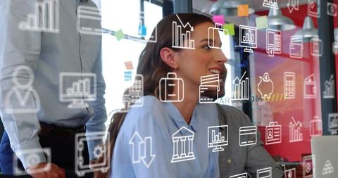 Seated woman reviewing laptop while team collaborating with digital business icons on glass