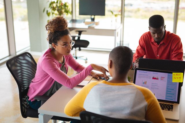 Multiracial team collaborating in a modern office environment, discussing project details while using laptops. Ideal for depicting teamwork, professional collaboration, and a diverse workplace in business presentations, corporate websites, and marketing materials.
