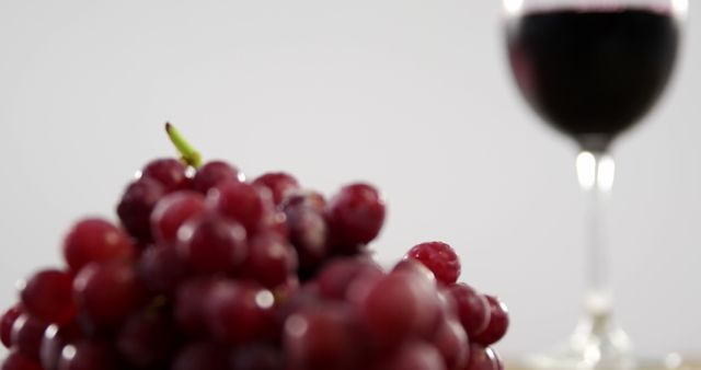 A bunch of red grapes is in focus in the foreground, with a glass of red wine blurred in the background, with copy space. Grapes and wine often symbolize abundance and celebration in various cultures.
