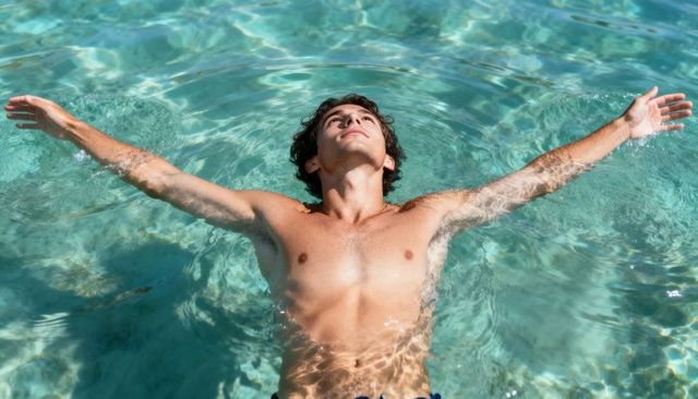 Sunlit young man floating in turquoise water arms outstretched relaxing