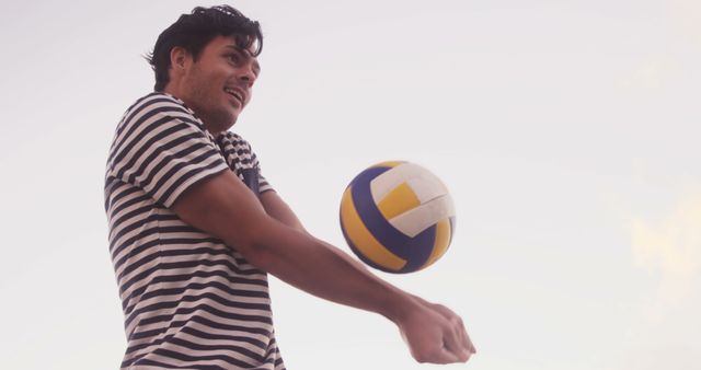Young man wearing striped shirt playing beach volleyball during sunset. Perfect for sports and outdoor activity related content, promotions for fitness and active lifestyle, or travel and leisure advertisements.