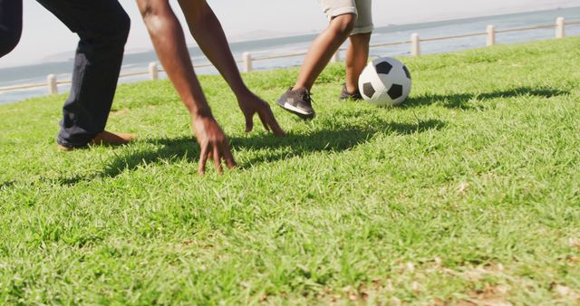 Children are playing soccer outdoors on a lush green lawn near a body of water. The focus is on their lower bodies and the soccer ball as they enjoy a sunny day. Perfect for use in advertisements promoting outdoor activities, sports gear, family fun, or recreational locations.