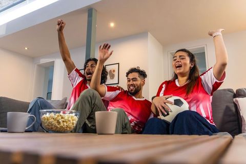 Friends cheering while watching soccer match at home wearing red jerseys holding popcorn
