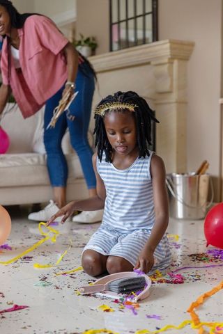 African american girl kneeling picking confetti while mom tidying living room after party