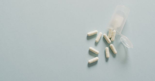 White capsules are spilling from a small plastic container onto a light blue surface. This image is ideal for use in medical, healthcare, or pharmaceutical advertisements, websites, and educational materials. It can also be utilized in blog posts or articles discussing medication, supplements, and health topics.