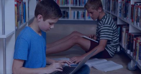 Two boys studying with laptop and tablet on library floor focusing on homework