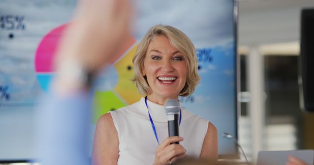 Businesswoman giving a confident presentation while holding a microphone and smiling at audience. Ideal for showcasing leadership, professional speaking engagements, corporate events, or motivational talks. Useful for articles, blogs, and marketing materials related to business, seminars, and leadership skills.