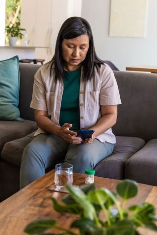 Mature woman using smartphone looking thoughtful with medication and glass of water