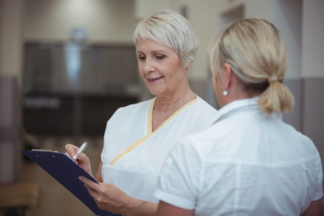 Two nurses are discussing patient records in a hospital corridor. One nurse is holding a clipboard and writing notes while the other listens attentively. This image can be used for healthcare-related content, medical teamwork, patient care, and hospital environment themes.
