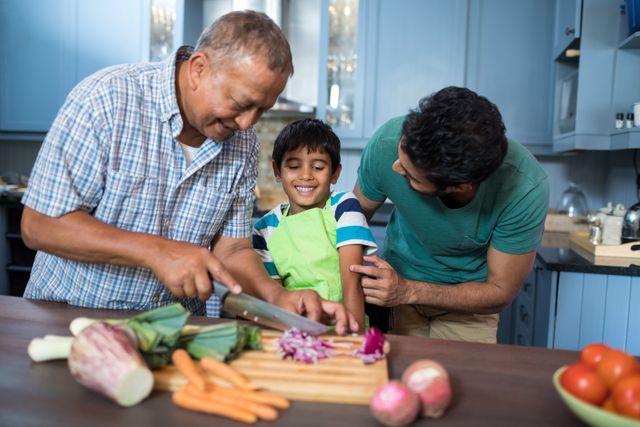 Grandfather cutting vegetables while father and son watch in a home kitchen. The scene captures a moment of family bonding and togetherness. Ideal for use in advertisements or articles about family life, healthy eating, and multigenerational activities.