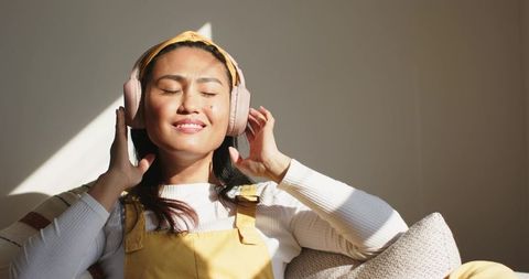 Asian woman relaxing and listening to music on couch with pink headphones mustard overalls