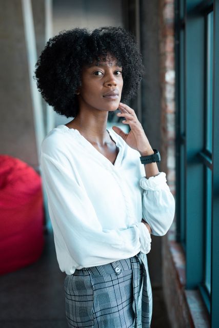 This image features a confident African American woman standing by a window in a cafe, looking at the camera. She is dressed in a white blouse and plaid pants, exuding a modern and stylish vibe. This image can be used for articles or advertisements focusing on urban lifestyle, fashion, independence, and modern women. It is also suitable for use in blogs or social media posts about confidence, personal style, and city living.