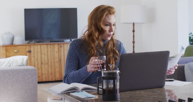 The image shows a woman with red hair in a cozy blue sweater working from home. She is seated at a desk with a laptop and holding a cup of coffee in one hand while looking at a piece of paper in the other hand. A French press and an open notebook are visible on the desk. A TV and cabinet are in the background. This image may be used to represent remote work, freelancing, studying, productivity, or home office environments.