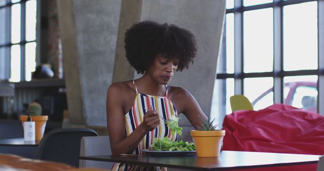 Young woman with curly hair enjoying a healthy salad at modern café with natural lighting. She wears a striped dress, adding chic vibrancy. Ideal for illustrating concepts of healthy lifestyle, casual dining, relaxed atmosphere, urban living, and wellness.