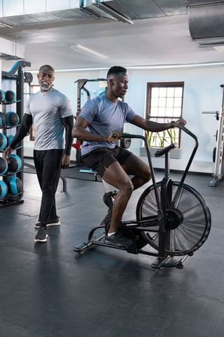 African american personal trainer coaching man pedaling air bike in modern fitness gym
