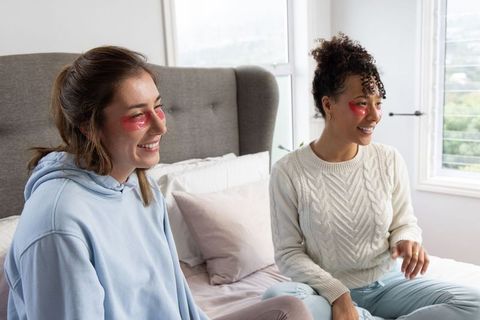 Diverse female friends sharing cozy skincare moment on bed with red under-eye gel patches