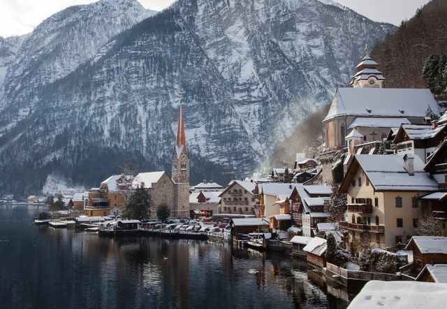 Picture shows idyllic winter scene of a snow-covered alpine village by a serene lake surrounded by mountains. Prominent features include charming houses, a tall church spire, and a peaceful atmosphere. Perfect for travel brochures, holiday cards, or winter destinations promotion.