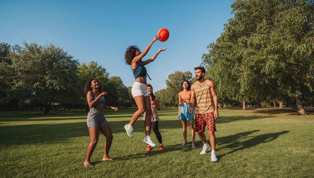 Group of friends playing frisbee in sunny park, woman jumping to catch orange disc outdoors