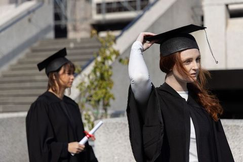 Two female graduates in black gowns on campus stairs adjusting caps and holding diploma