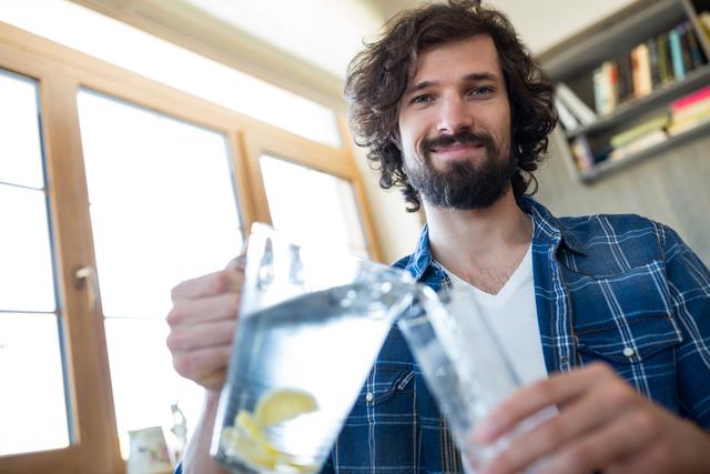 A man with a beard and casual attire smiling as he pours lemonade into a glass. Suitable for use in advertisements promoting healthy lifestyles, beverage brands, home living concepts, and relaxation themes.