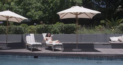 Sunlit poolside relaxing woman reading on striped lounge under umbrella in private garden
