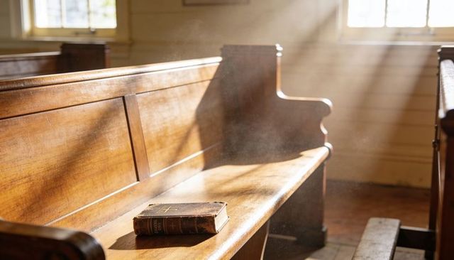 Sunlight streaming through chapel window onto wooden pew holding worn leather bible and dust motes