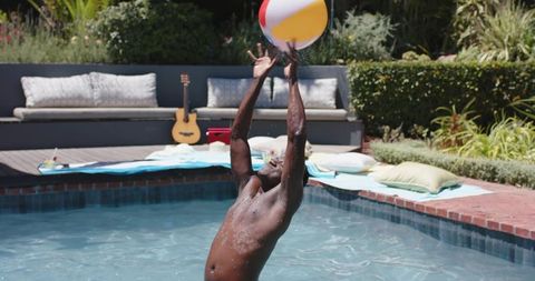 African american man reaching for beach ball while rising from backyard pool on sunny day