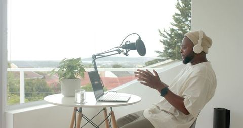 African american man recording podcast on sunny balcony with laptop and boom microphone