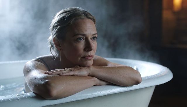 Middle-aged woman resting on enamel tub rim, gazing contemplatively amid steam and mist
