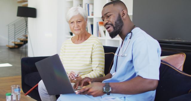 Nurse in blue scrubs assisting elderly woman with laptop usage in a cozy home environment. Useful for illustrating home healthcare services, senior support, technology aid for seniors, and professional caregiving.